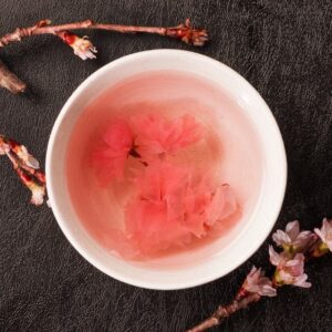 A bowl of pale pink liquid with pink flowers floating in it sits on a dark textured surface, surrounded by blossoming branches.
