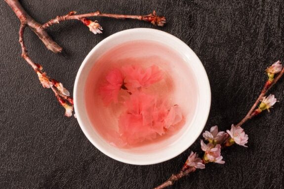 A bowl of pale pink liquid with pink flowers floating in it sits on a dark textured surface, surrounded by blossoming branches.