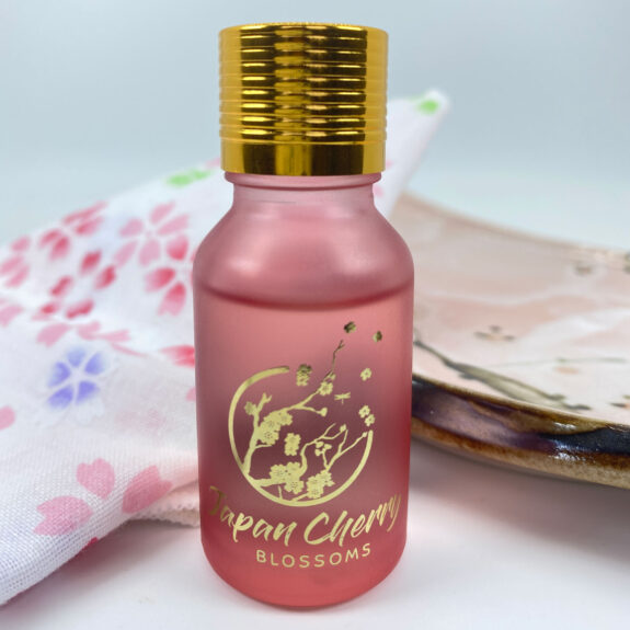 A pink frosted bottle with gold cap and cherry blossom design sits next to a patterned cloth and plate.