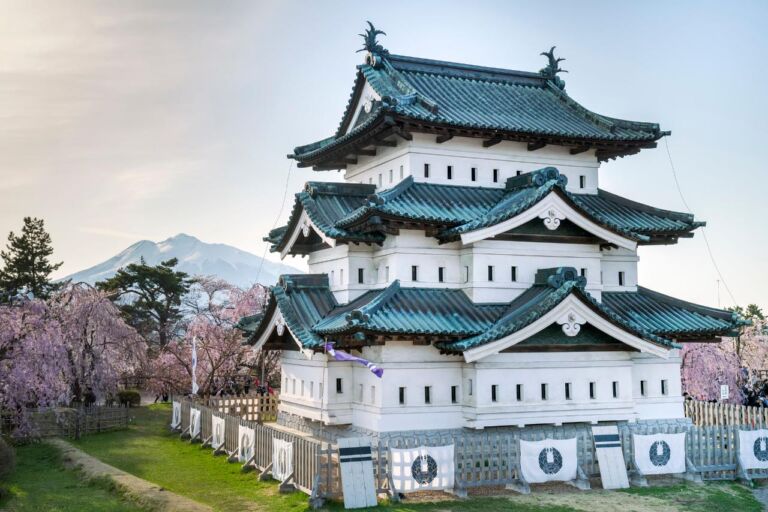 A stunning view of a traditional Japanese castle with white walls and teal-green tiled roofs, surrounded by blooming cherry blossoms, with a mountain in the background.