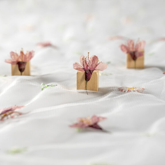 A serene close-up of delicate pink flowers held in small wooden blocks, scattered on a white, sheer fabric.