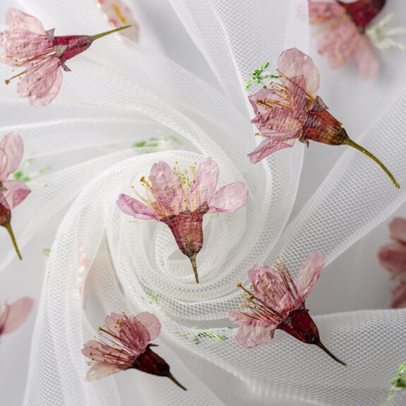 An overhead shot of delicate pink cherry blossom-like flowers scattered on a swirling, sheer white mesh fabric, creating a soft, dreamy effect.
