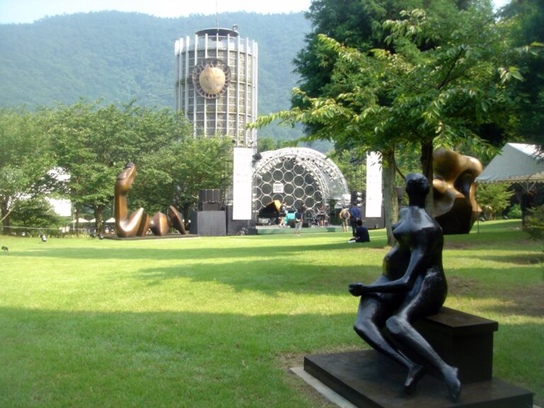 A grassy park with sculptures, a stage, and a tall tower with a sun symbol in the background.