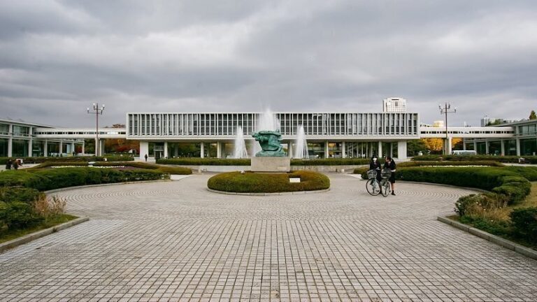 The Hiroshima Peace Memorial Park features a central sculpture with fountains in a circular space, framed by a modern building and symmetrical gardens under a cloudy sky.