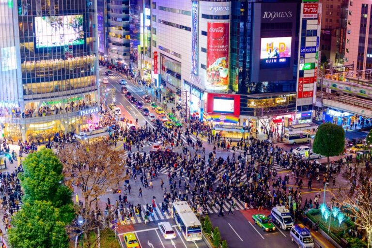 This is a nighttime aerial view of the Shibuya Crossing in Tokyo, showing a large crowd of pedestrians crossing the street amidst bright lights and buildings with illuminated billboards.