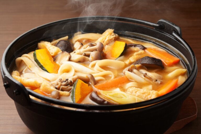 A steaming hot pot of Japanese udon noodle soup filled with mushrooms, carrots, pumpkin, tofu, and leeks sits on a wooden table.