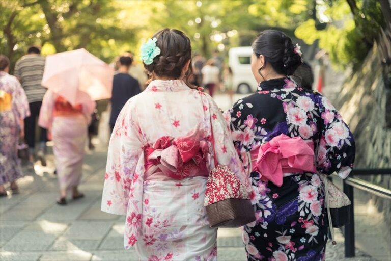 Two women wearing colorful kimonos and obis stroll down a tree-lined path, showcasing traditional Japanese attire with floral patterns and intricate hair accessories.