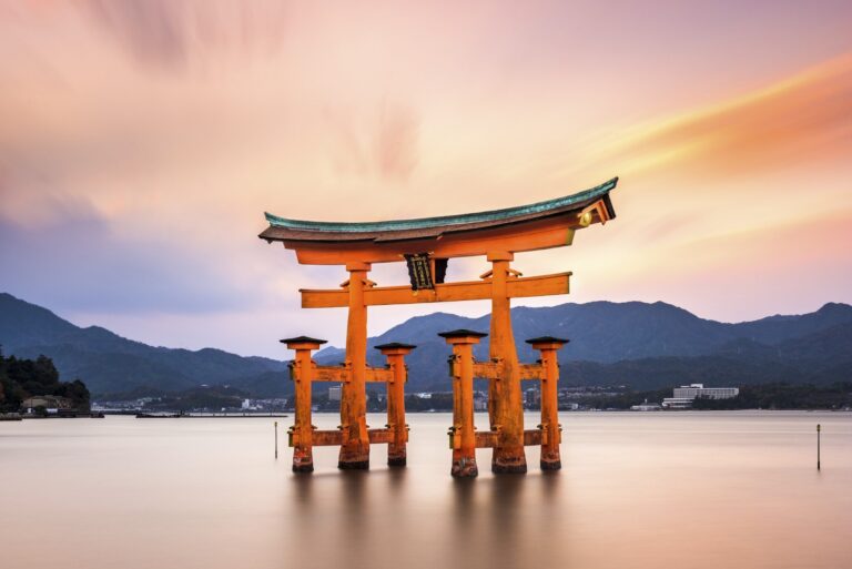The Itsukushima Shrine torii gate stands majestically in the water at sunset, with mountains in the background, creating a serene and picturesque scene.